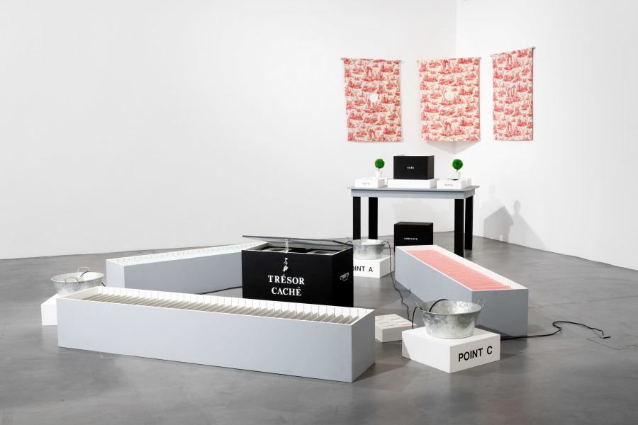 Renée Green, Mise-en-Scène, 1991, Installation. Mixed media: table, painted wooden structures, 117 rubber-stamped painted wood plaques, vinyl type, 79 documents (color photographs, b/w photographs, postcards) magnifying glass, cotton gloves, three metal basins, three mechanical elements, three mounted postcards on wood, three hanging textiles with collage, two topiaries, sound. Courtesy Frac des Pays de la Loire, Carquefou. Photo: Flavio Karrer.