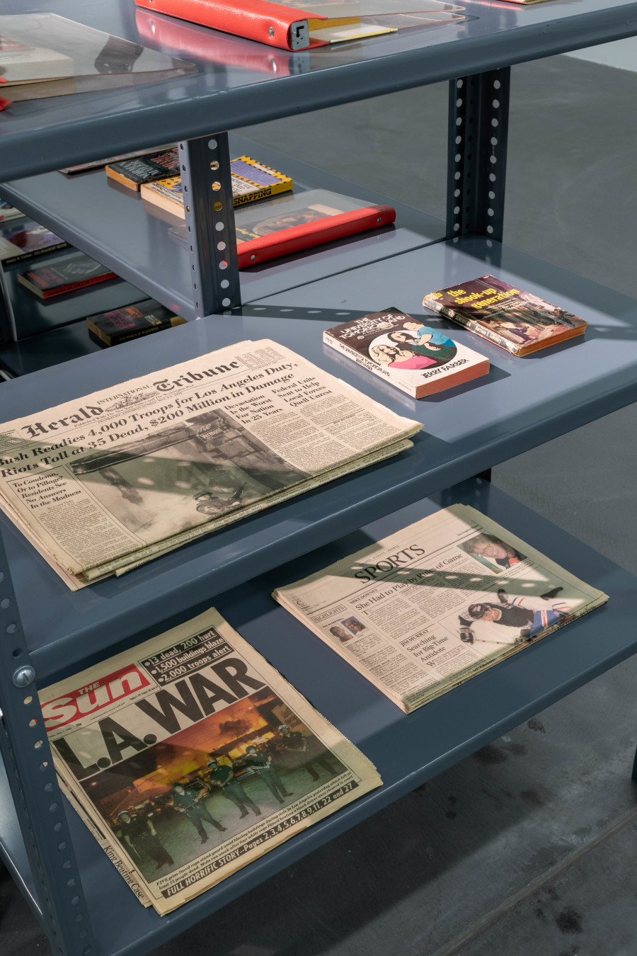 Renée Green, Import/Export Funk Office, 1992 - 1993, Installation. Mixed media: metal shelf structure, books, magazines, newspapers, ephemera, BETA video cases, 25 hours of digitized video, two monitors, one video projection, cassette cases, sound, acrylic signs [COLLECTANEA, IMPORT/EXPORT FUNK OFFICE, FUNK STATION (1-4)], four wooden structures [Funk Stations] acrylic signs on four shelves with four cassette players [LEXICON; LOCAL (EAST?); LOCAL (WEST?); IMPORT], 30 wooden plaques with rubber-stamped text [LEXICON]. Courtesy The Museum of Contemporary Art, Los Angeles. Gift of Gaby and Wilhelm Schürmann. Photo: Flavio Karrer.