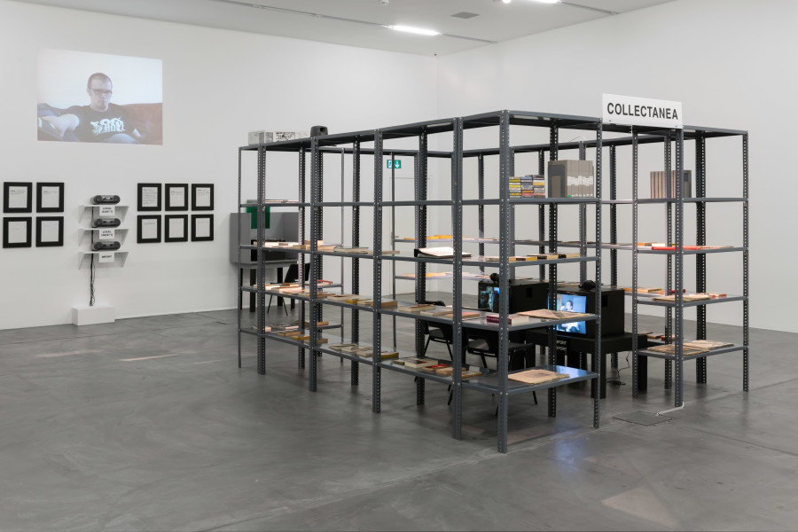 Renée Green, Import/Export Funk Office, 1992 - 1993, Installation. Mixed media: metal shelf structure, books, magazines, newspapers, ephemera, BETA video cases, 25 hours of digitized video, two monitors, one video projection, cassette cases, sound, acrylic signs [COLLECTANEA, IMPORT/EXPORT FUNK OFFICE, FUNK STATION (1-4)], four wooden structures [Funk Stations] acrylic signs on four shelves with four cassette players [LEXICON; LOCAL (EAST?); LOCAL (WEST?); IMPORT], 30 wooden plaques with rubber-stamped text [LEXICON]. Courtesy The Museum of Contemporary Art, Los Angeles. Gift of Gaby and Wilhelm Schürmann. Photo: Flavio Karrer.