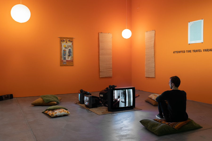 Renée Green, Übertragen/Transfer, 1997, Installation. Mixed media: 16 color photographs, six b/w photographs, three lithographs, vinyl type on wall (Attempted Time Travel Variable Sites Continuing Circuit “Complex Fate”), five videos (Partially Buried, 1996; Conversation with Janice and Lonnie “Lumumba” Turner, 1996; Übertragen/Transfer (Samir Alschausky, Henrietta Schroeter, Simin Farkhondeh), 1996; Super 8 Footage, 1996; Underground, 1976, a film by Emile de Antonio, Mary Lampson, and Haskell Wexler), sound, one hanging appliqué, two paper ball lamps, two window roller shutters, sisal carpet, pillows, wall treatment. Courtesy Museum Ludwig, Köln / Ankauf mit Unterstützung der Ludwig Stiftung 2001. Photo: Flavio Karrer.