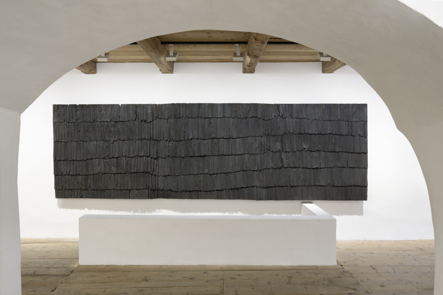 Exhibition view, Augustas Serapinas, Roof of a House from Steponių Village, 2022, charred reclaimed wooden shingle roof, 185 x 625 x 24 cm. Photo: Ralph Feiner, Courtesy of the artist and Galerie Tschudi